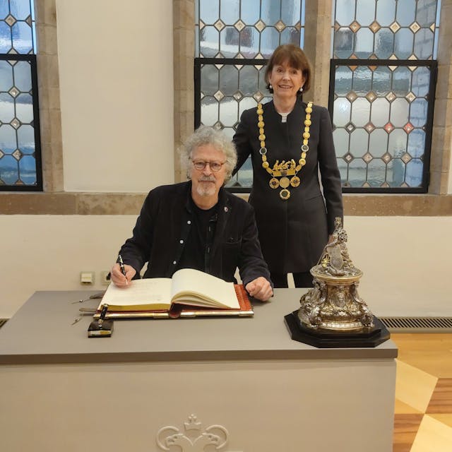 Wolfgang Niedecken sitzt an einem Tisch und schreibt in das Goldene Buch, hinter ihm steht Oberbürgermeisterin Henriette Reker.