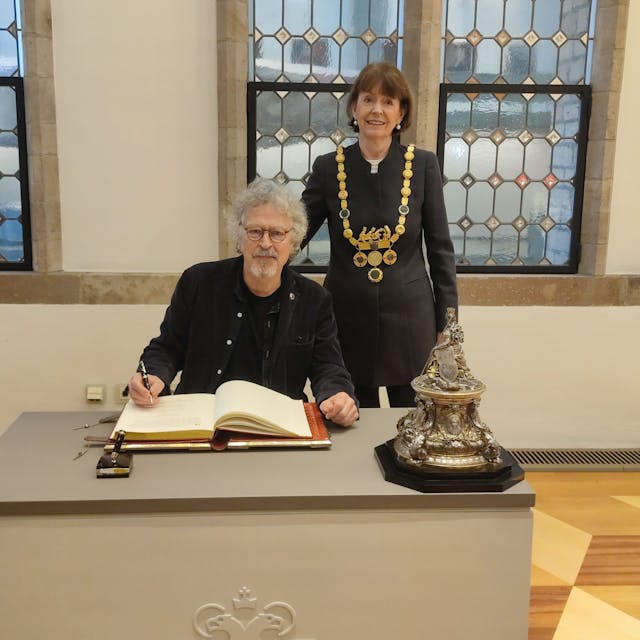 Wolfgang Niedecken sitzt an einem Tisch und schreibt in das Goldene Buch, hinter ihm steht Oberbürgermeisterin Henriette Reker.