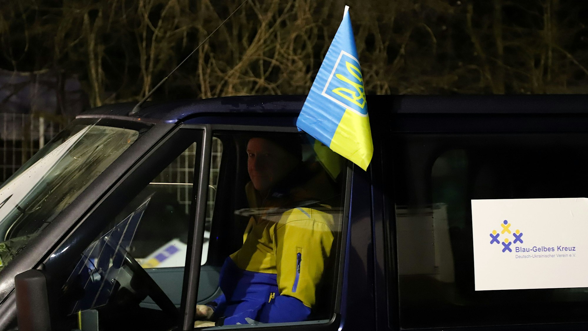 Ein Mann in blau-gelber Jacke sitzt in einem Transporter mit der Aufschrift „Blau-Gelbes Kreuz“ und einer Ukraineflagge.