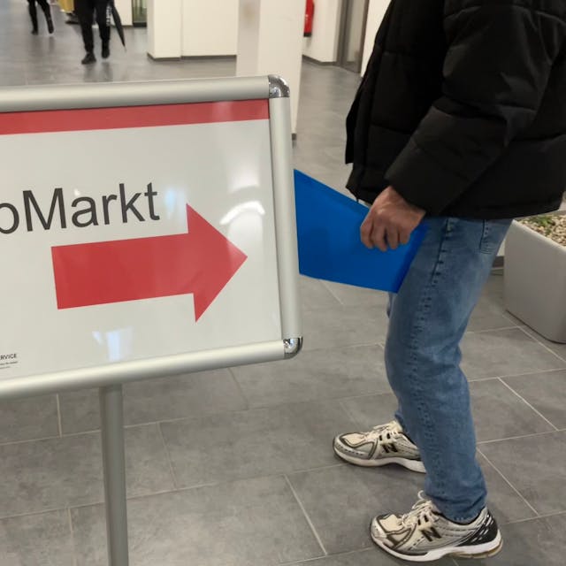 In der Agentur für Arbeit in Köln weist ein Schild auf den Jobmarkt für Geflüchtete hin. Ein Mann mit einer Bewerbungsmappe folgt dem Hinweispfeil.