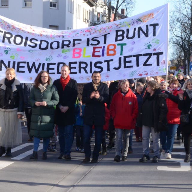 Ein Protestzug, in der ersten Reihe gehen Männer und Frauen unter einem Transparent. Darauf steht "Troisdorf ist bunt" und "Nie wieder ist jetzt"