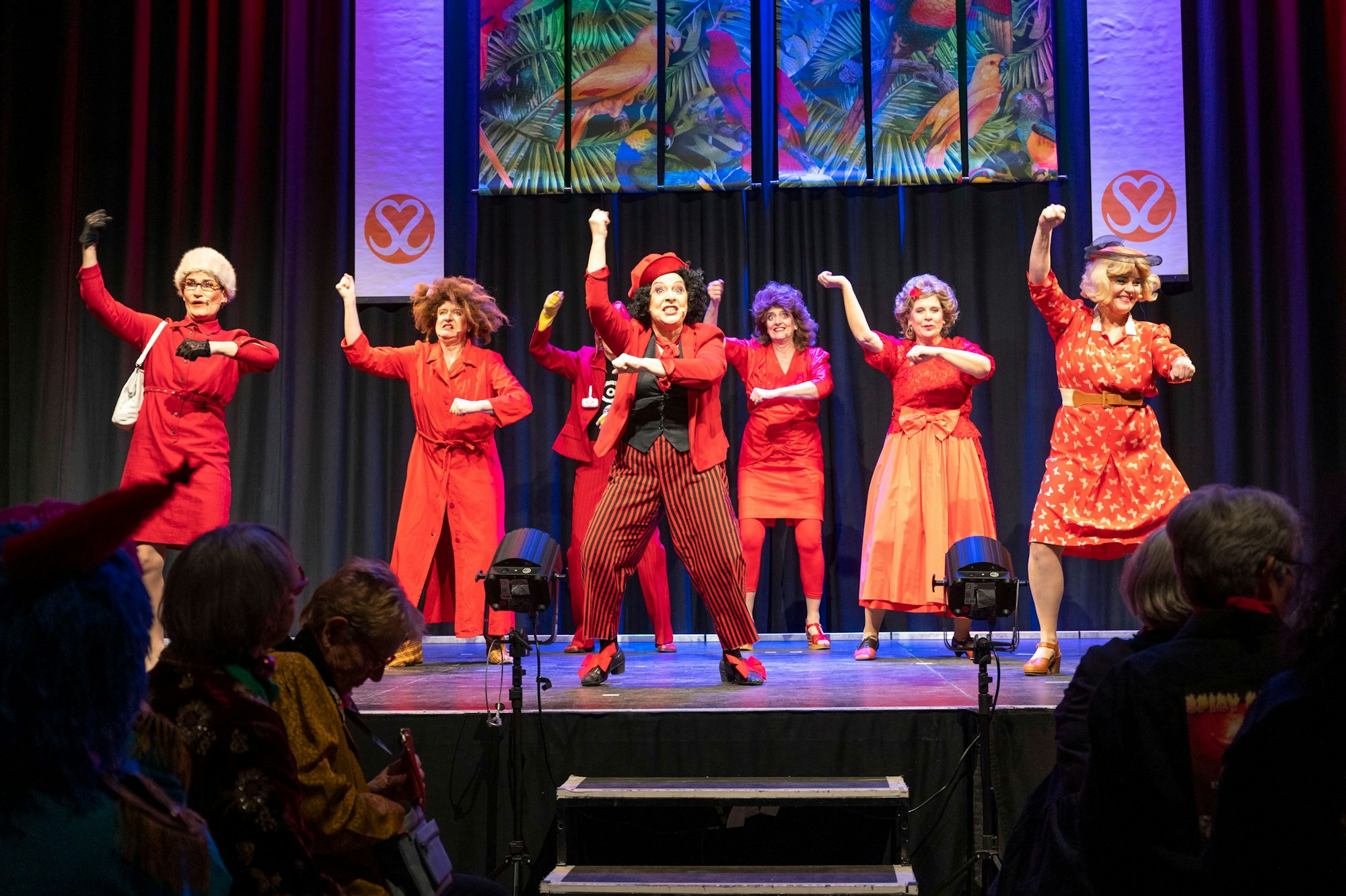Sieben Frauen in roten Kostümen tanzen auf der Bühne.