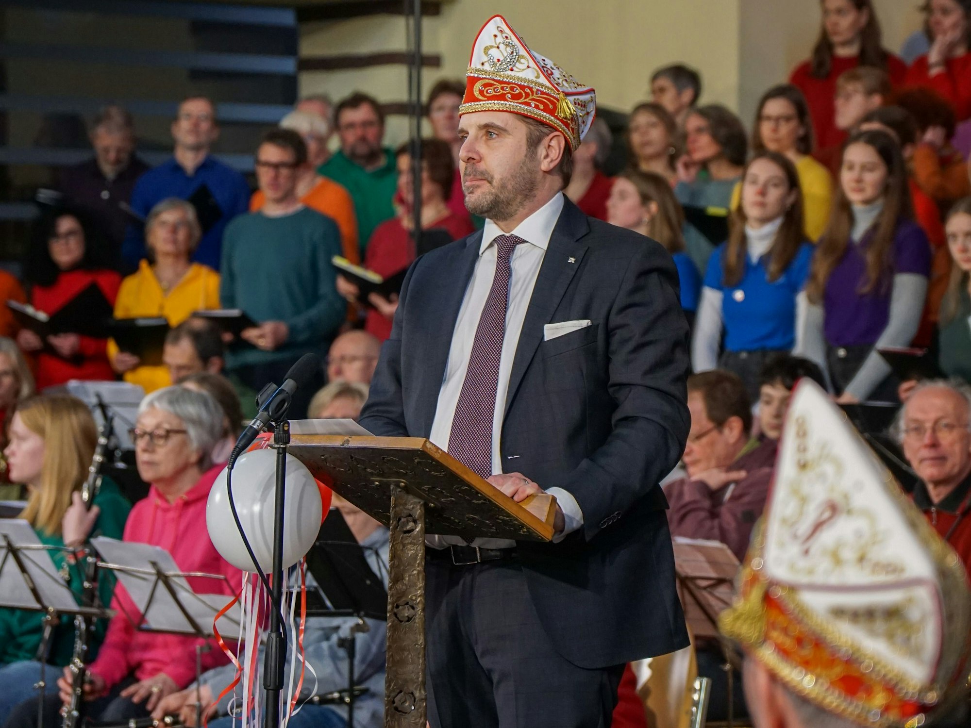 Ein Mann im Anzug mit Narrenkappe auf dem Kopf, steht an einem Rednerpult, hinter ihm sind Musiker und Chormitglieder zu sehen.