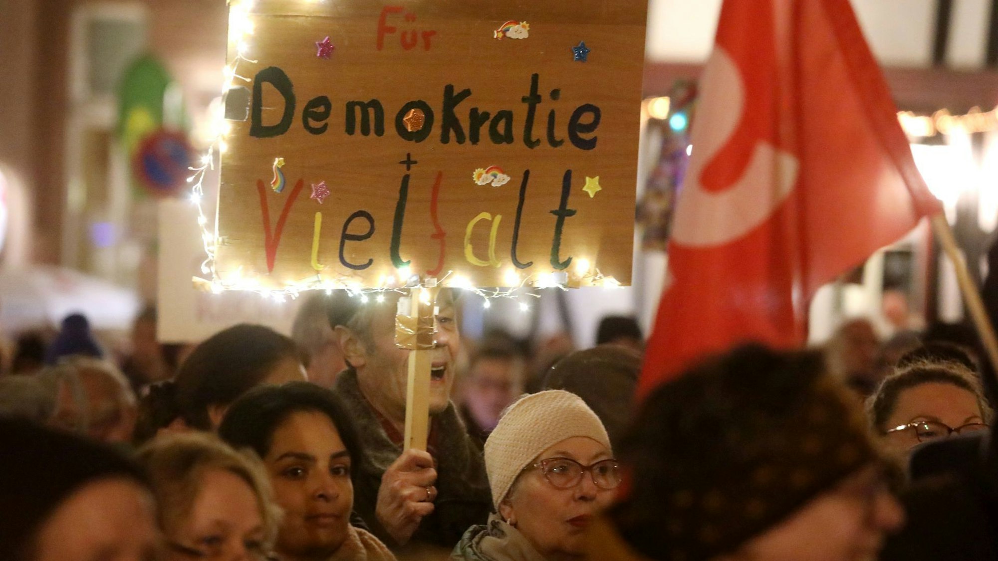 Teilnehmer einer Kundgebung halten Fahnen und Protestschilder. „Für Demokratie und Vielfalt“ steht auf einem bunten Schild mit Lichterkette.