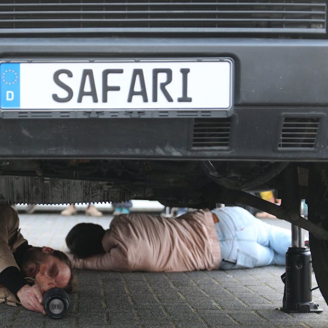 Auf dem Foto sind zwei Künstler zu sehen, die auf der Straße unter einem Fahrzeug liegen.