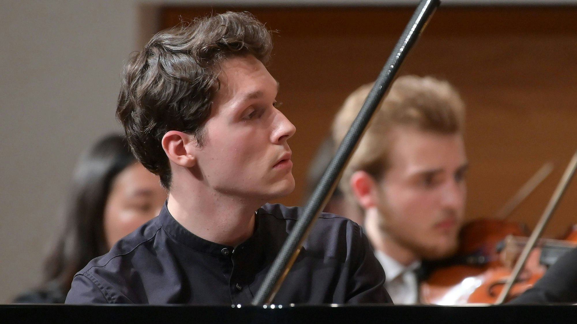 Das Bild zeigt den Pianisten Lukas Sternath 2023 bei einem Konzert des Orchesters der Angelika Prokopp Sommerakademie der Wiener Philharmoniker. Er sitzt am Klavier und schaut auf die rechte Bildseite, sein Gesicht ist im Profil zu sehen. Im Hintergrund zwei Streicher.