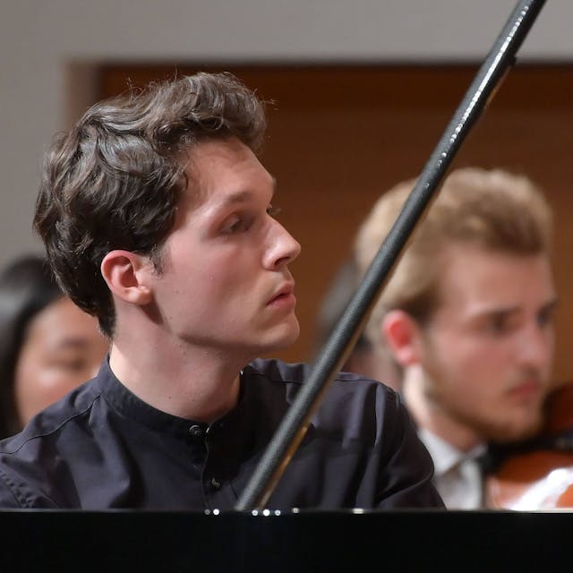 Das Bild zeigt den Pianisten Lukas Sternath 2023 bei einem Konzert des Orchesters der Angelika Prokopp Sommerakademie der Wiener Philharmoniker. Er sitzt am Klavier und schaut auf die rechte Bildseite, sein Gesicht ist im Profil zu sehen. Im Hintergrund zwei Streicher.