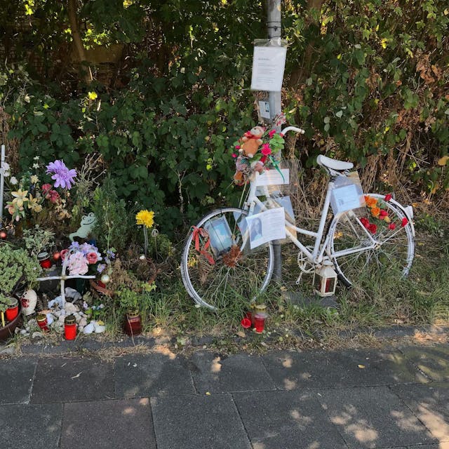 Ein weißes Fahrrad, Blumen und Kerzen stehen am Wegesrand.