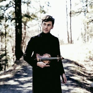 Violinist Benjamin Beilman steht auf einem Weg im Wald. Er hält seine Violine und trägt schwarze Kleidung.&nbsp;