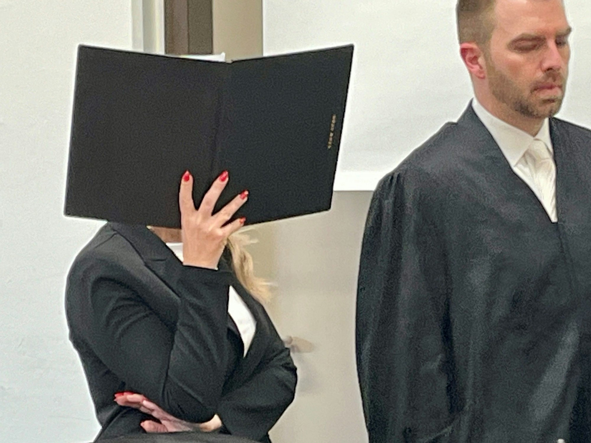 Eine Frau hält sich eine Mappe vor das Gesicht, neben ihr steht ein Anwalt.