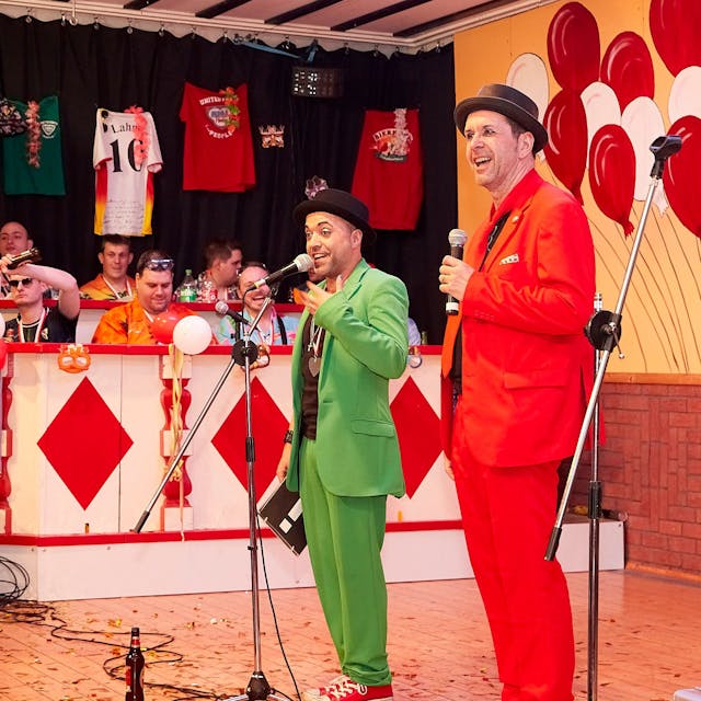 Zwei Redner, einer in rotem Anzug, einer in grünem Anzug, stehen auf einer Karnevalsbühne vor Mikrofonen.