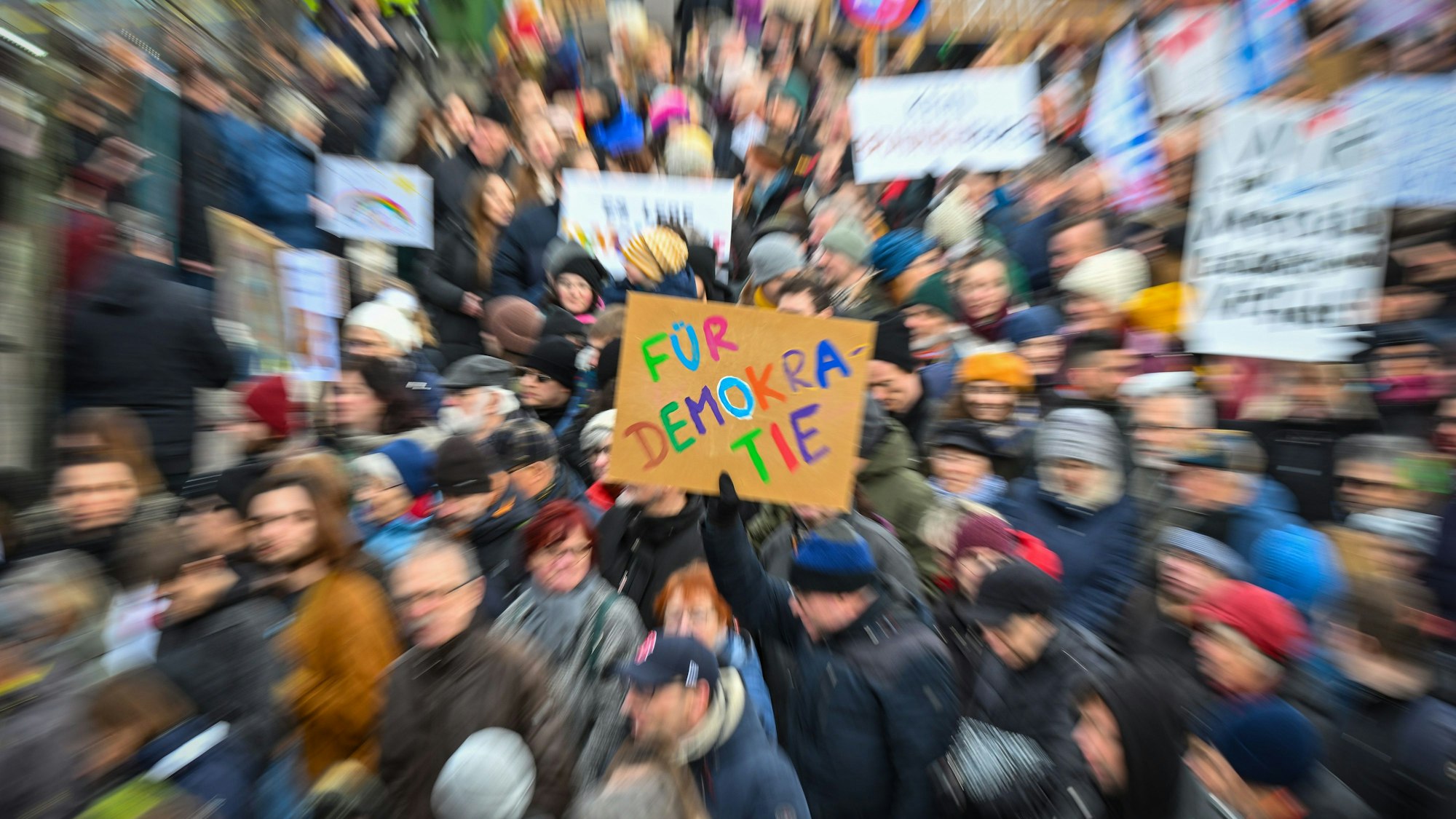 Das Bild zeigt Teilnehmer einer Demonstration, die Plakate hochhalten. Während alle Personen und Schilder verzerrt dargestellt sind, ist nur ein Plakat in der Mitte des Fotos scharf abgebildet. Auf ihm ist zu lesen „Für Demokratie“.