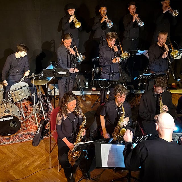 Die Musikerinnen und Musiker stehen und sitzen in der klassischen Big-Band-Aufstellung auf der Bühne, vorne fünf Akteure mit Saxofon.
