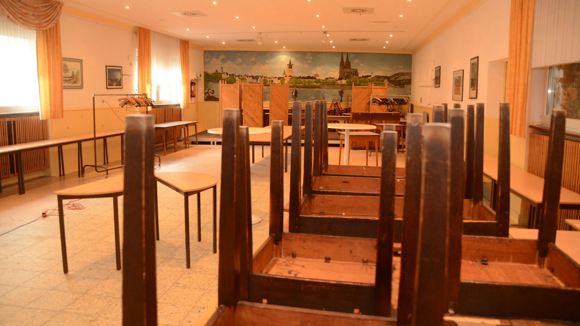 Der Saal einer Gaststätte: An der Wand ein gemaltes Panorama der Stadt Köln, rechts aufeinander gestellte Tische.
