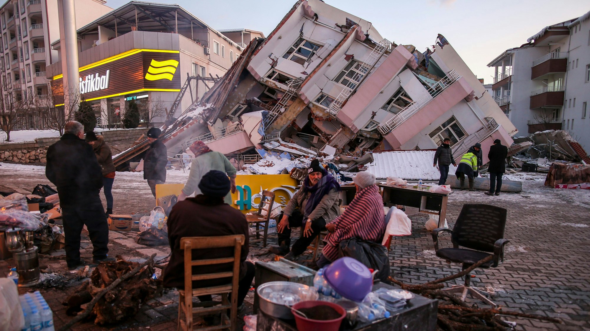 Im türkischen Ort Golbasi in der Provinz Adiyaman sitzen im Februar 2023 Menschen auf Stühlen auf der Straße. Im Hintergrund ist ein eingestürztes Gebäude zu sehen.