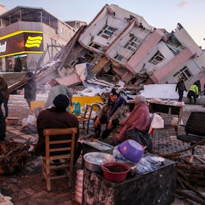 Im türkischen Ort Golbasi in der Provinz Adiyaman sitzen im Februar 2023 Menschen auf Stühlen auf der Straße. Im Hintergrund ist ein eingestürztes Gebäude zu sehen.
