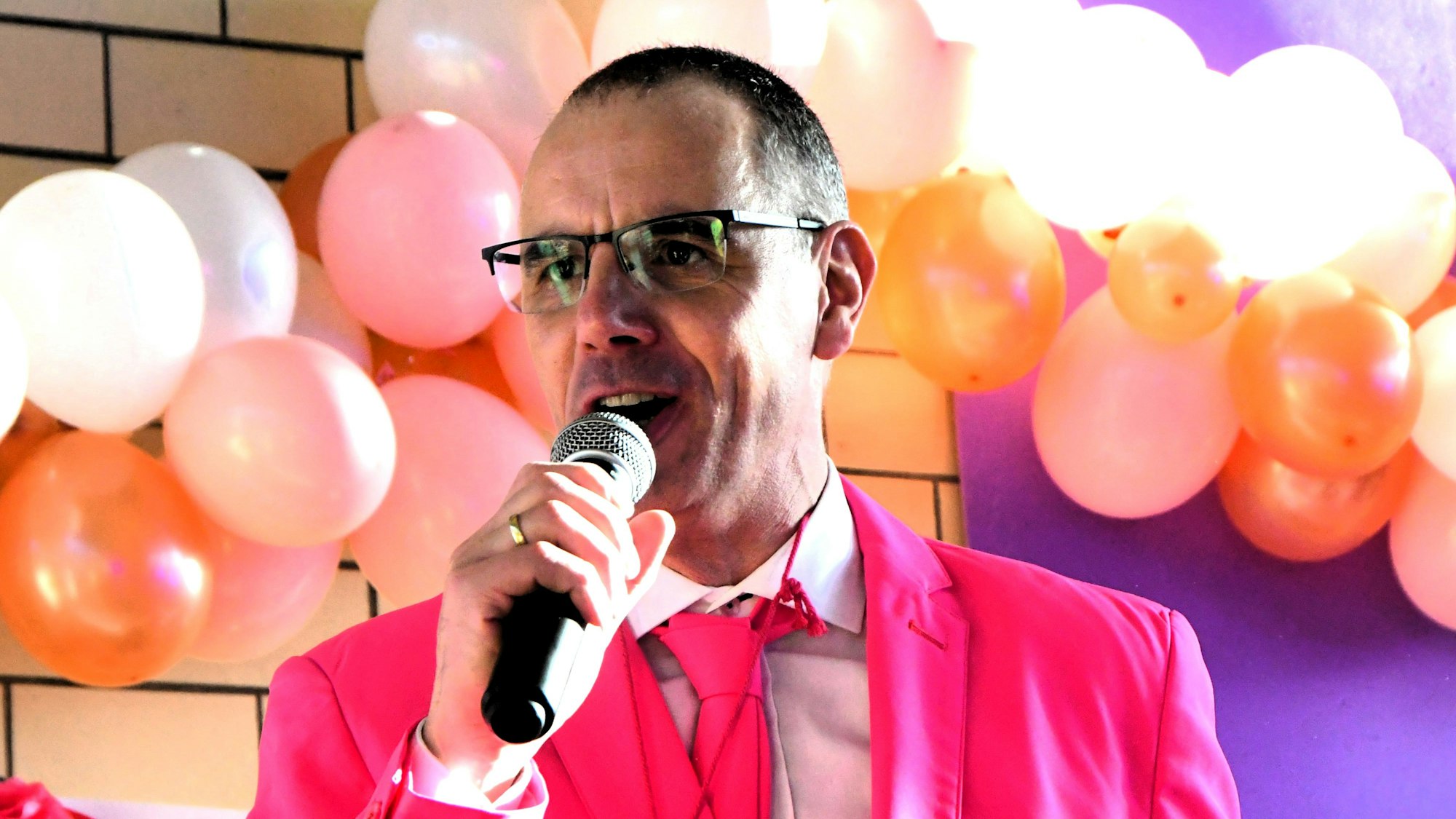 Werner Haag, mit Anzug und Krawatte in Pink, spricht auf der mit Ballons dekorierten Bühne ins Mikrofon.