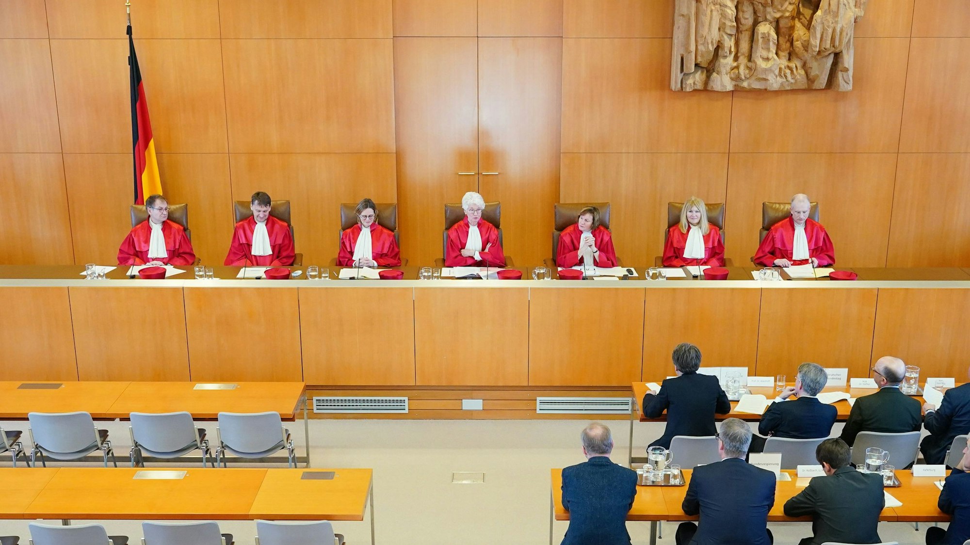Sieben Verfassungsrichterinnen und -richter in roten Umhängen sitzen in Karlsruhe während einer Verhandlung nebeneinander und blicken in den Saal.
