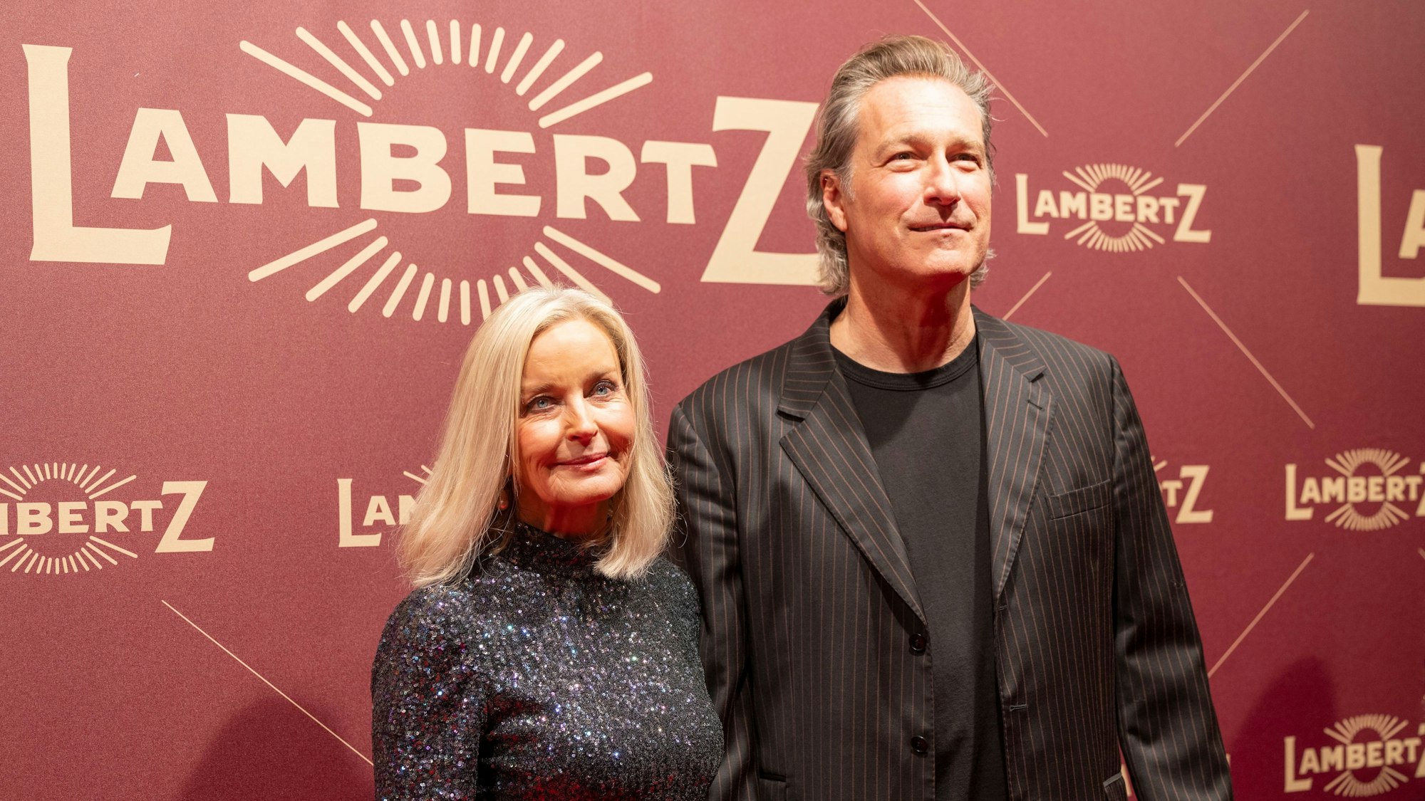 John Corbett steht mit Partnerin Bo Derek vor einer roten Wand, auf der Lambertz steht.
