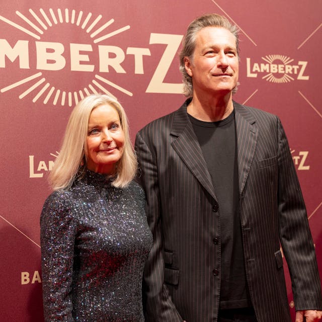 John Corbett steht mit Partnerin Bo Derek vor einer roten Wand, auf der Lambertz steht.
