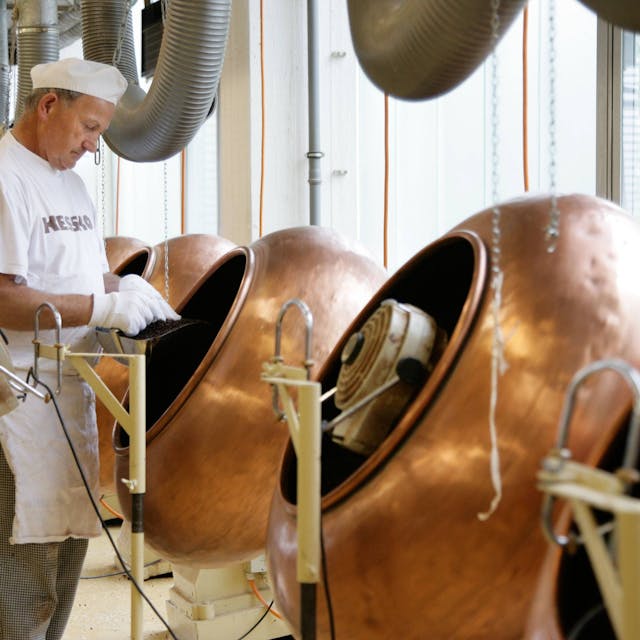 Ein Mann arbeitet an Maschinen, die zur Schokoladenherstellung dienen. Er steht links im Bild und trägt weiße Arbeitskleidung. Die Maschinen sind bronzefarben.