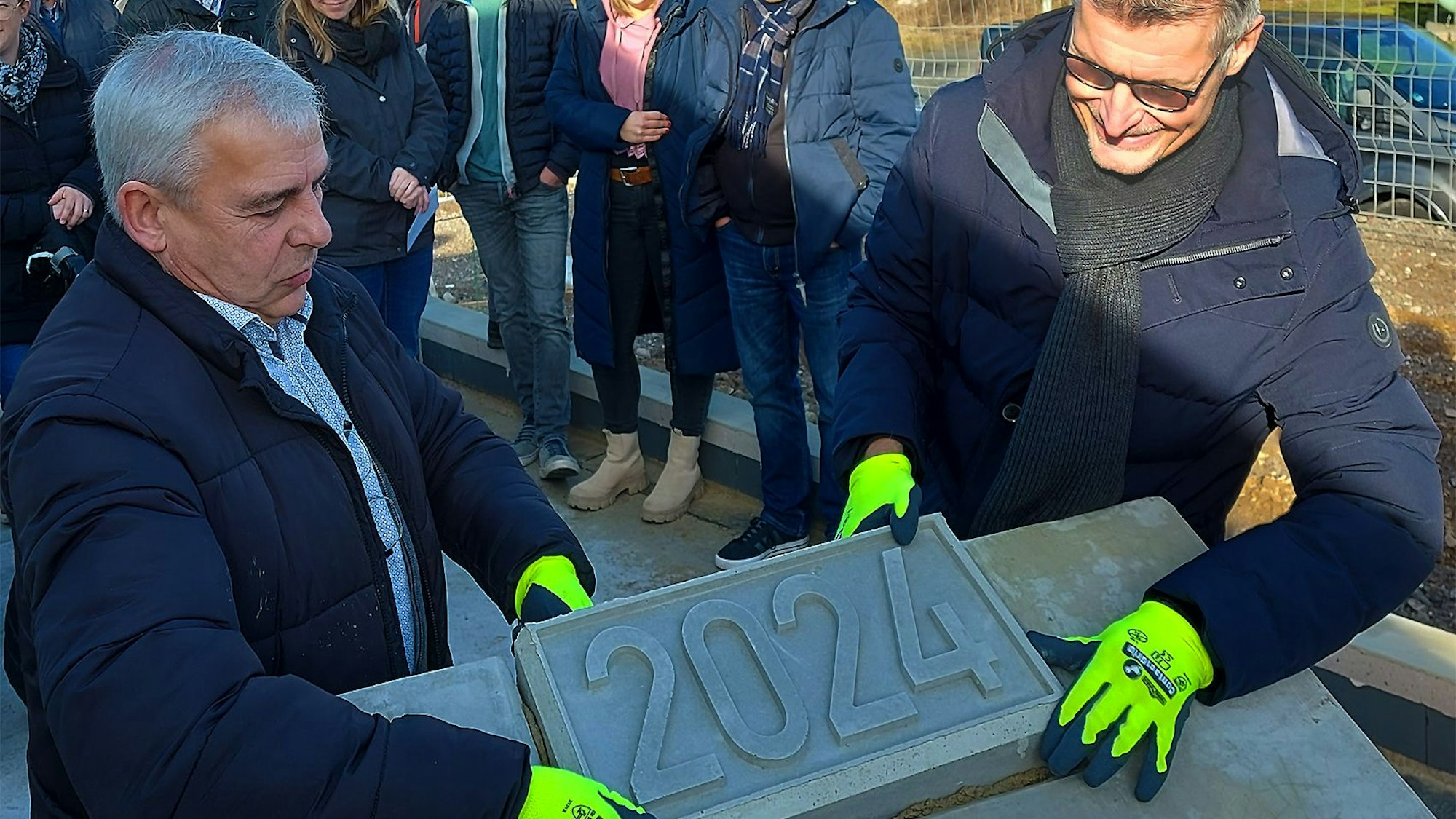 Zwei Männer, Hans-Josef Bohnen und Han Lembach, tragen gelbe Arbeitshandschuhe und halten einen Beton-Stein mit der Aufschrift „2024“ in den Händen. Sie setzen ihn in eine dafür vorgesehene Platte ein.
