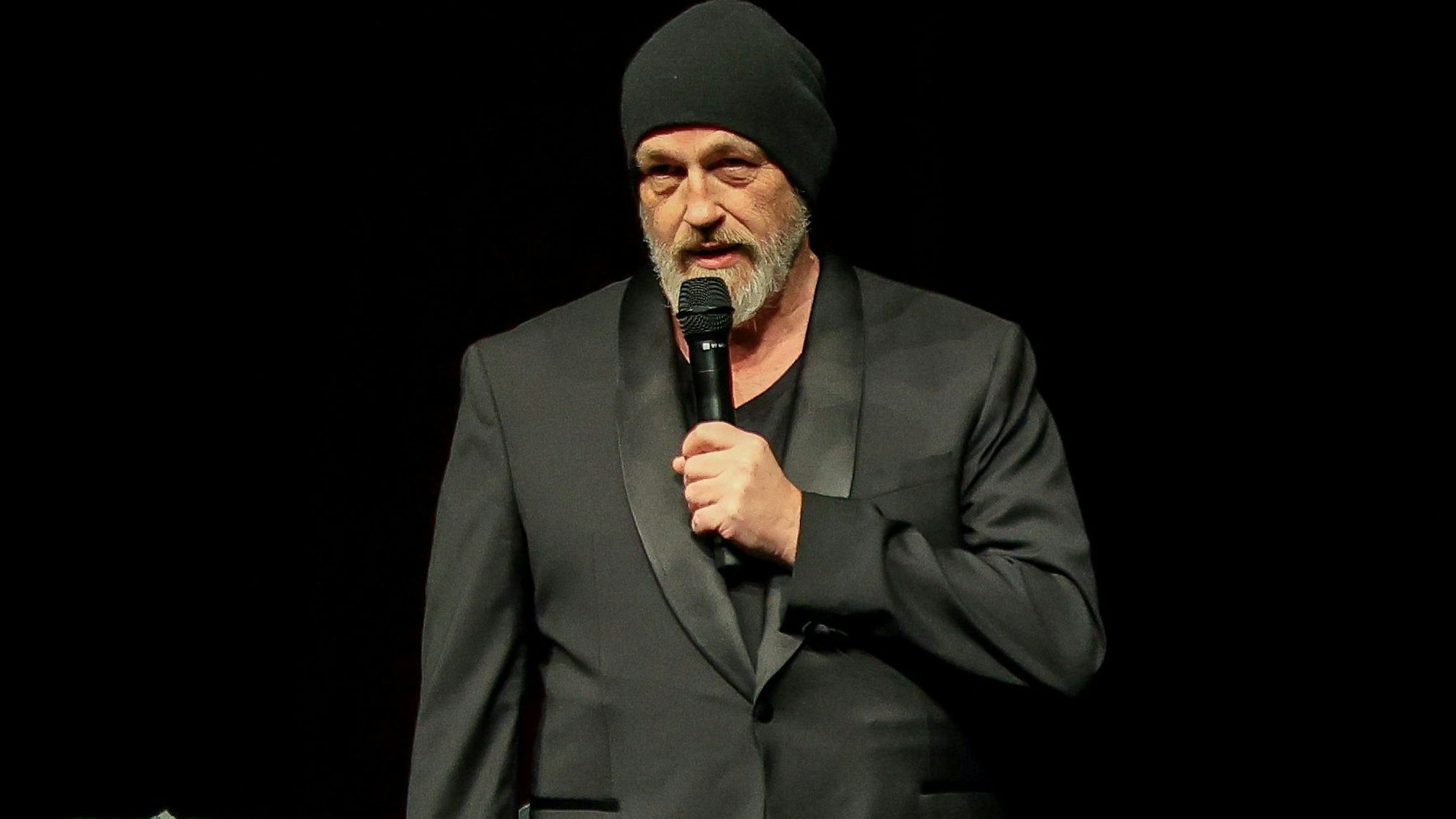 Ein Mann ganz in Schwarz gekleidet und mit einer schwarzen Mütze hält sich ein Mikrofon an den Mund.