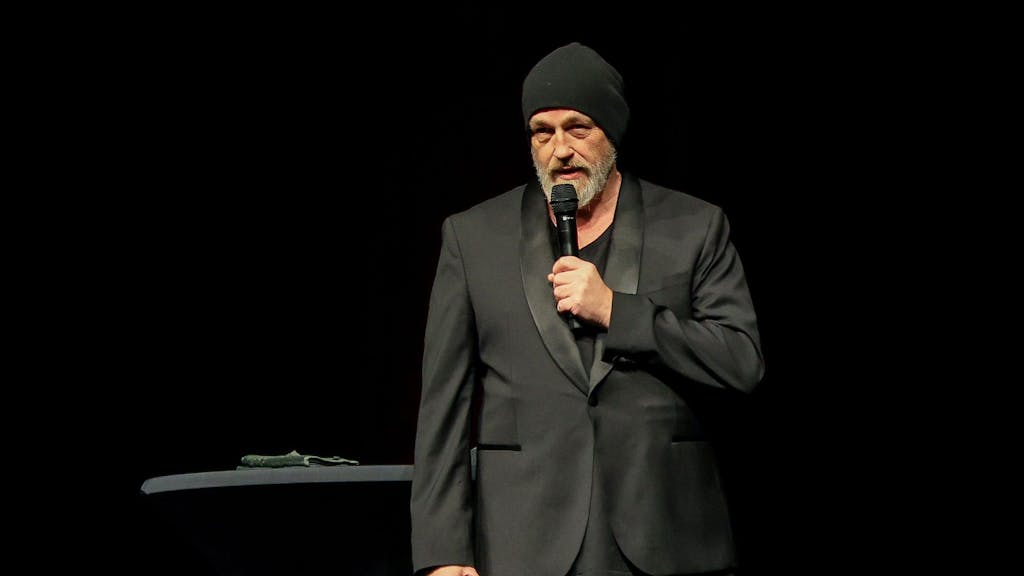 Ein Mann ganz in Schwarz gekleidet und mit einer schwarzen Mütze hält sich ein Mikrofon an den Mund.&nbsp;