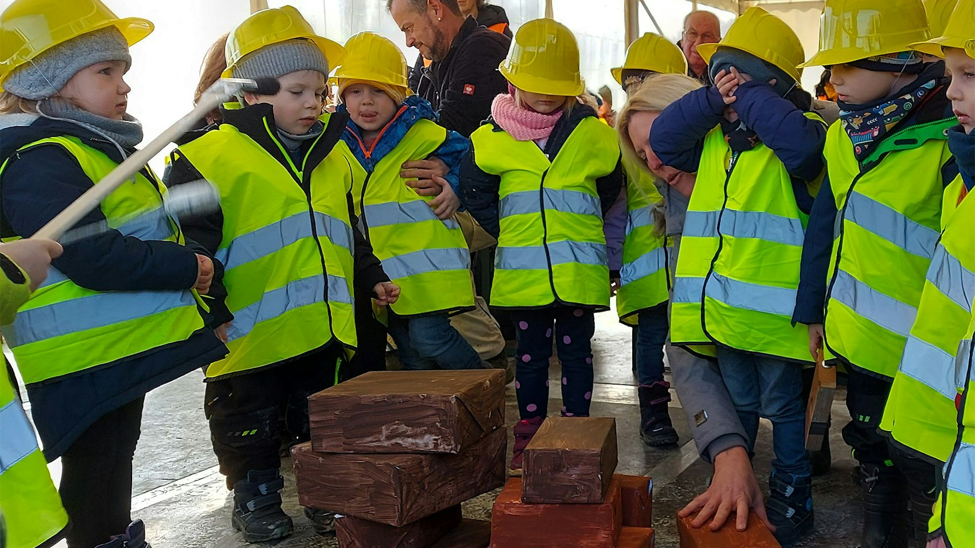 Einige Kinder stehen in gelben Warnwesten und mit gelben Helmen um ein paar Steine und spielen fleißige Handwerker.