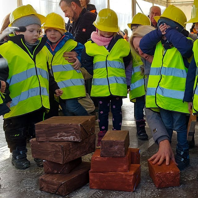 Einige Kinder stehen in gelben Warnwesten und mit gelben Helmen um ein paar Steine und spielen fleißige Handwerker.