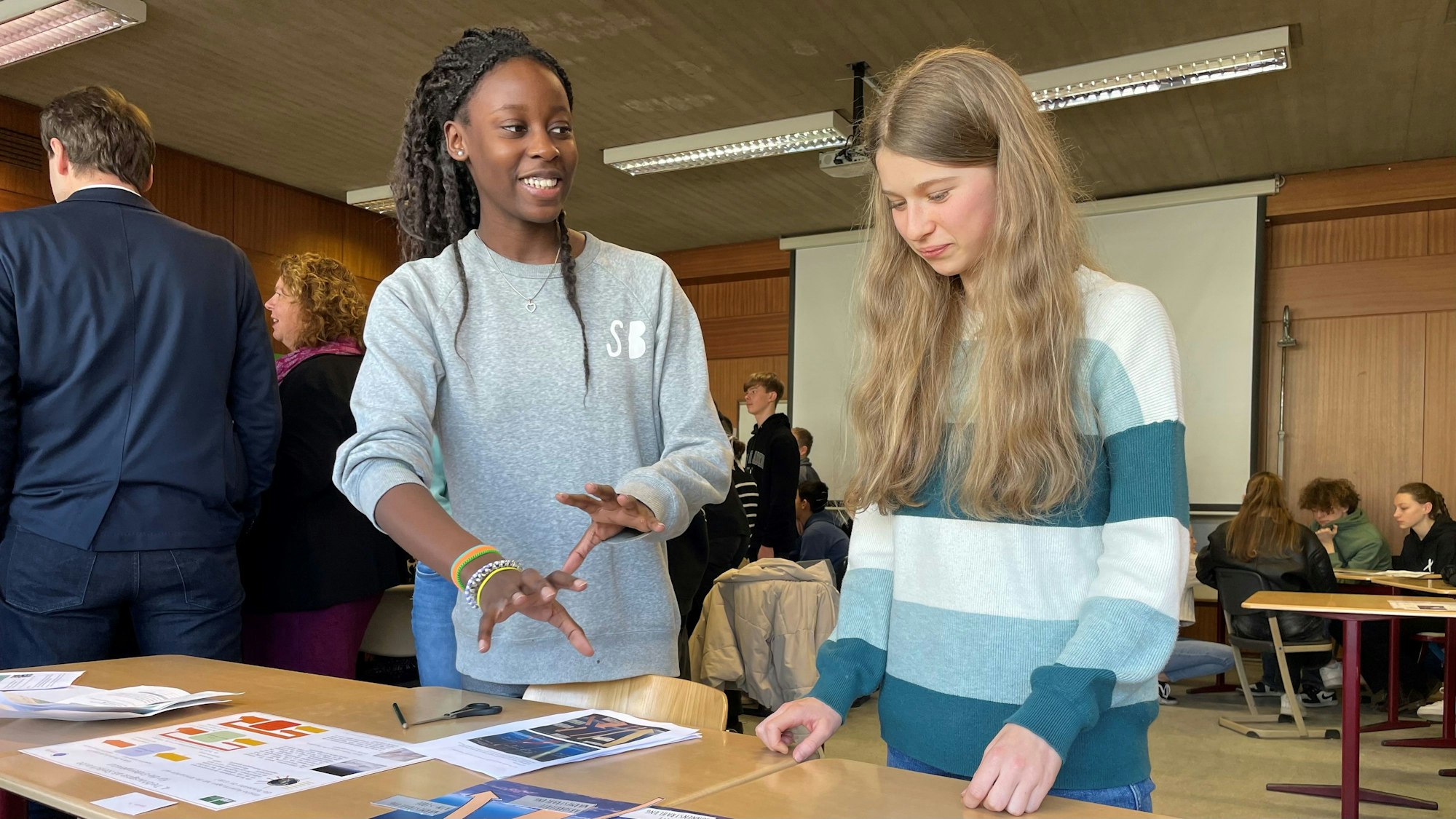 Zwei Schülerinnen der achten Klasse reden hinter einem Tisch stehen miteinander. Auf dem Tisch liegen Materialien aus dem Klimakoffer.