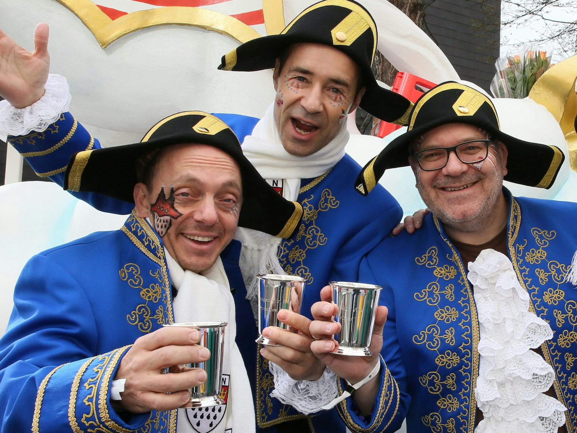 Drei kostümierte Männer stehen vor einem Karnevalswagen.