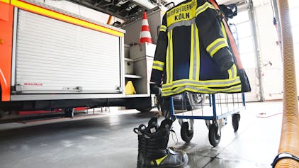 Eine Feuerwehruniform hängt über einem Haken, im Hintergrund steht eine Feuerwehrauto.