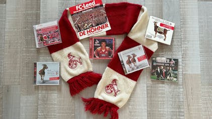 Auf einem rot-weißen Fan-Schal des 1. FC Köln sind verschiedene CDs sowie eine Schallplatte mit Liedern über den Verein drapiert.