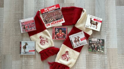 Auf einem rot-weißen Fan-Schal des 1. FC Köln sind verschiedene CDs sowie eine Schallplatte mit Liedern über den Verein drapiert.