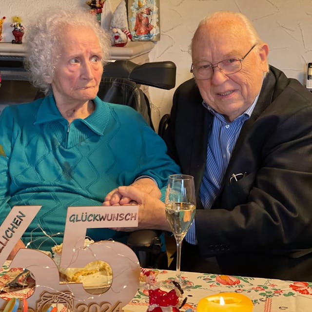 Hildegard und Horst Ziethen sitzen an einem Tisch, auf dem ein Strauß gelber und roter Rosen steht. Vor ihnen stehen Sektgläser.