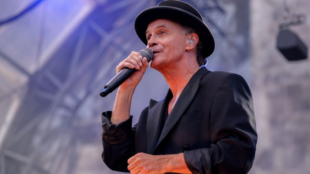 Peter Brings steht mit schwarzem Anzug und schwarzem Hut auf der Bühne und singt in ein Mikrofon.