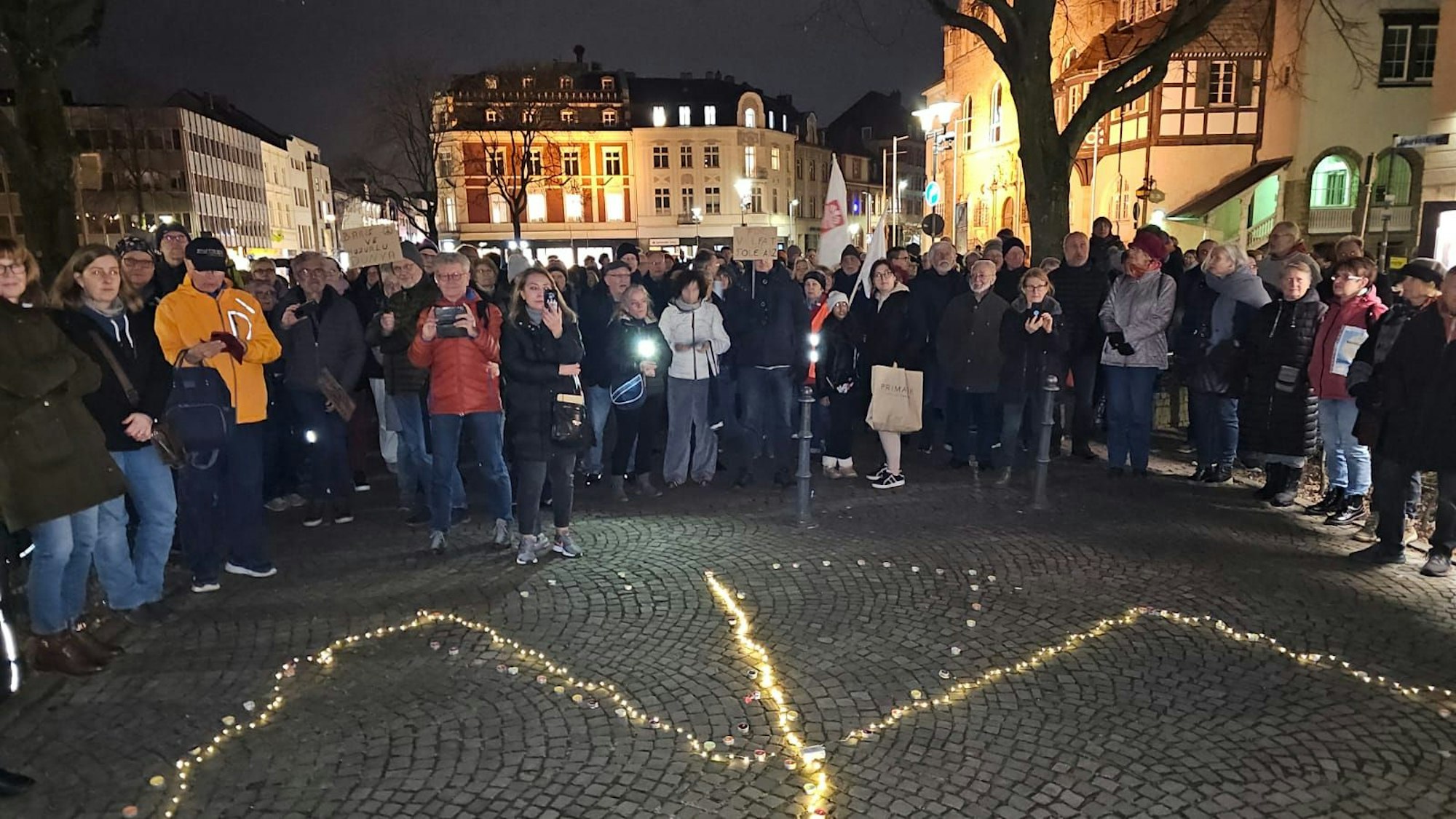 Menschen stehen auf dem Konrad-Adenauer-Platz von Bergisch Gladbach, auf dem Boden ist ein leuchtendes Peace-Zeichen zu sehen.