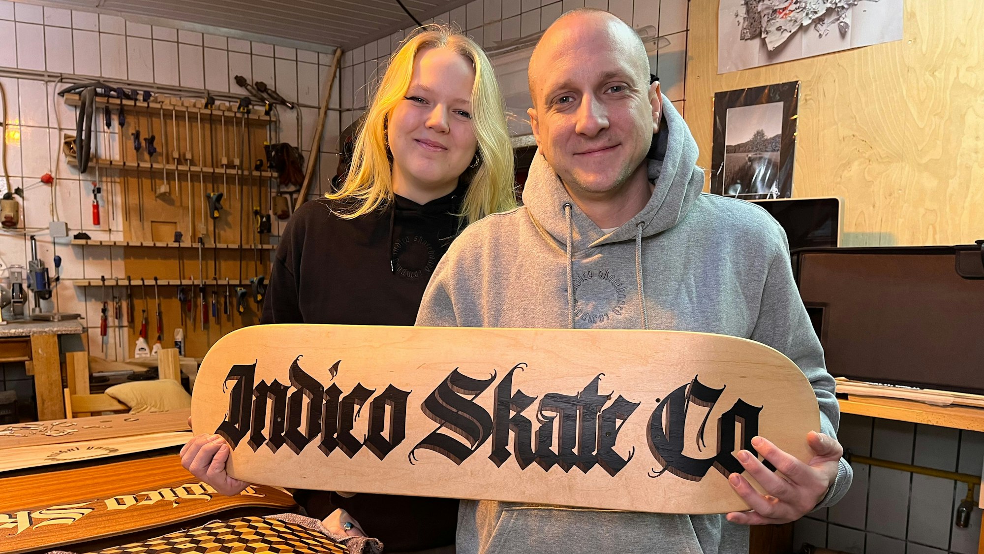 Eine junge Frau mit blondem Haar und ein Mann mit Glatze und Hoodie stehen nebeneinander. Er hält ein Holzstück, auf dem Indico Skate Co steht.