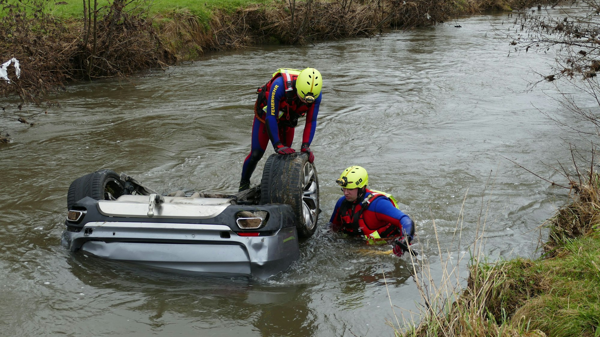 Zwei Männer in Neoprenanzügen stehen in einem Fluss an einem Auto, bei dem nur die Hinterräder aus dem Wasser ragen.