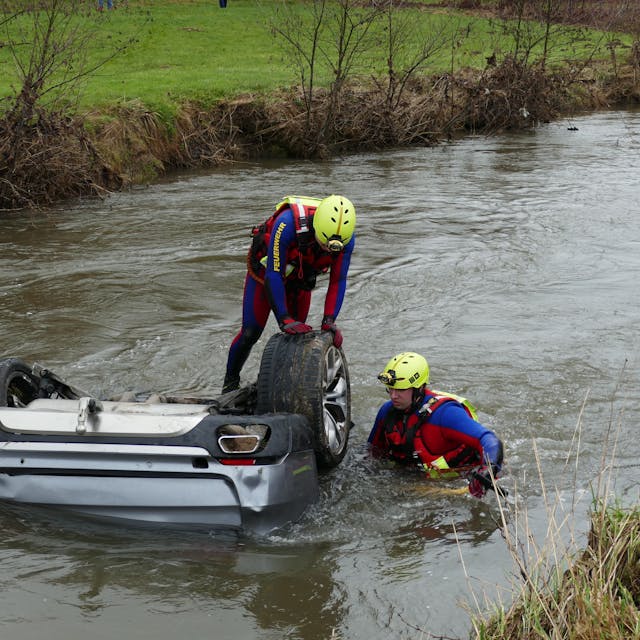 Zwei Männer in Neoprenanzügen stehen in einem Fluss an einem Auto, bei dem nur die Hinterräder aus dem Wasser ragen.