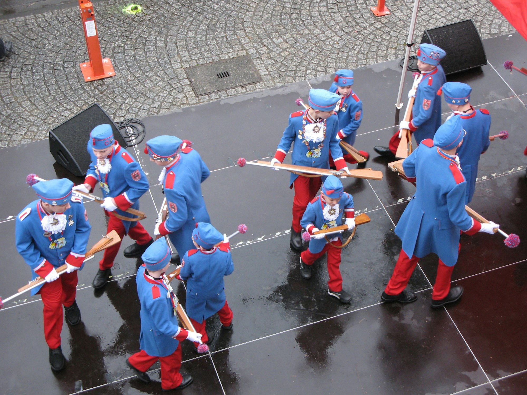 Kinder in blau-roten Stadtsoldaten-Uniformen in Tanzformation auf einer Bühne, von oben fotografiert.