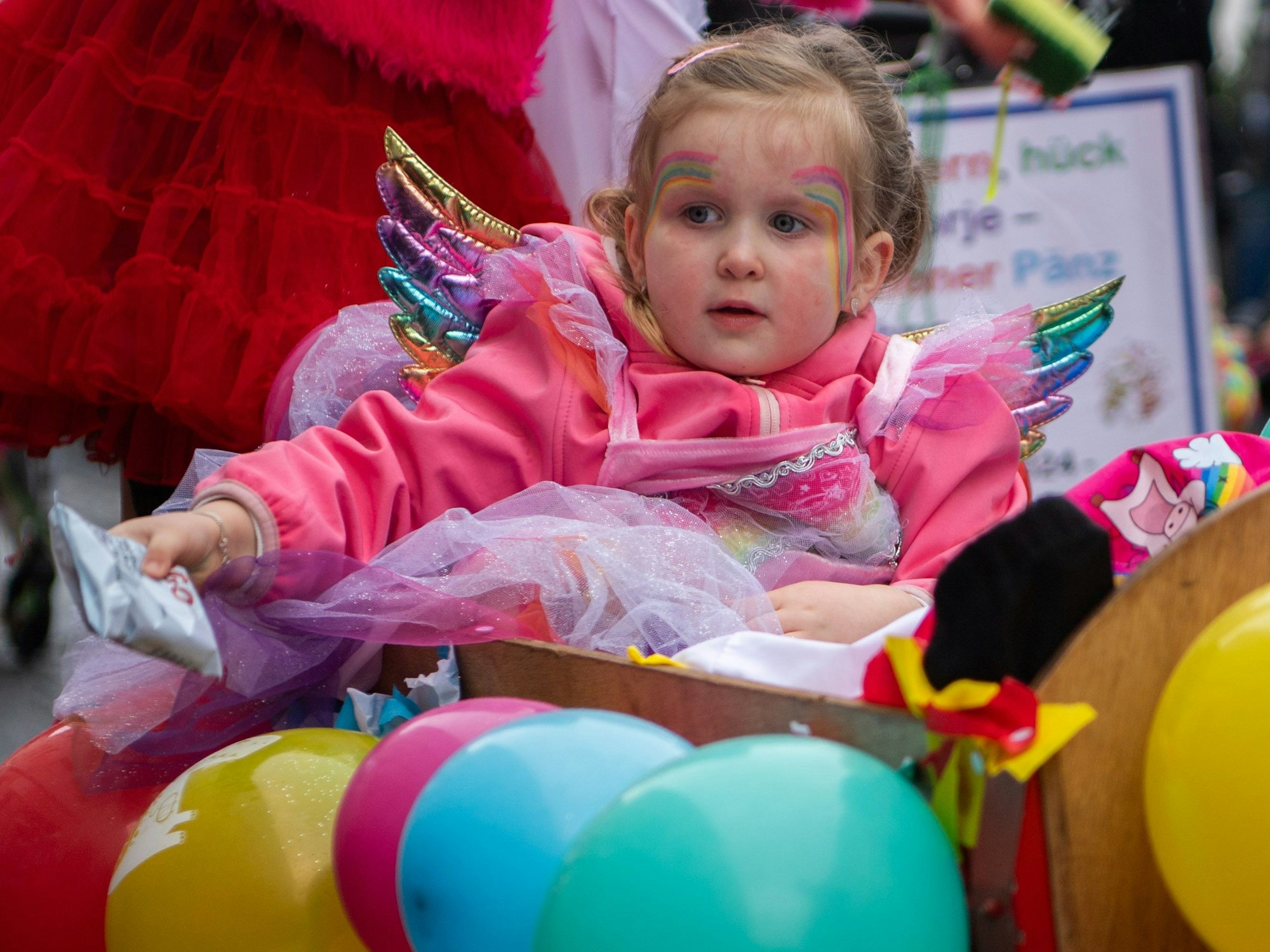 Das Bild zeigt ein Mädchen, das als Regenbogen kostümiert ist. Es sitzt in einem mit bunten Luftballons geschmückten Bollerwagen.