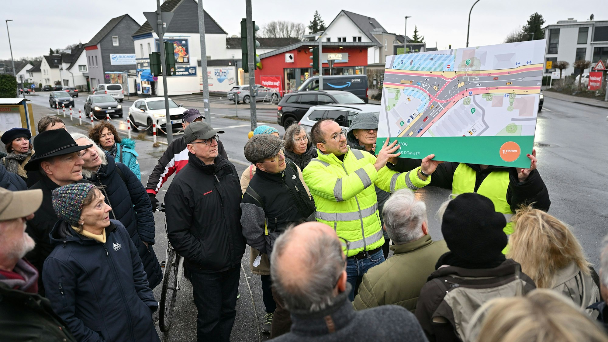 Zwei Planer halten ein Plakat hoch, das die vorgesehene Verkehrsführung zeigt. Um sie herum stehen Teilnehmer der Veranstaltung. Im Hintergrund sieht man die Straßenkreuzung.
