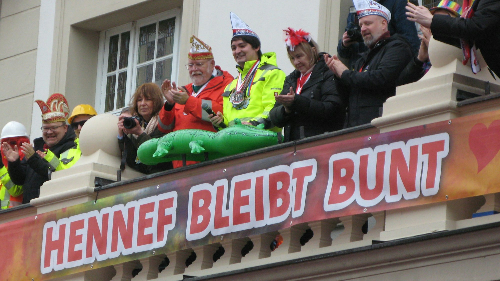 Mehrere applaudierende Personen auf einem Balkon, dabei der Hennefer Bürgermeister mit einem aufblasbaren Krokodil, davor das Transparent mit der Aufschrift „Hennef bleibt bunt“.