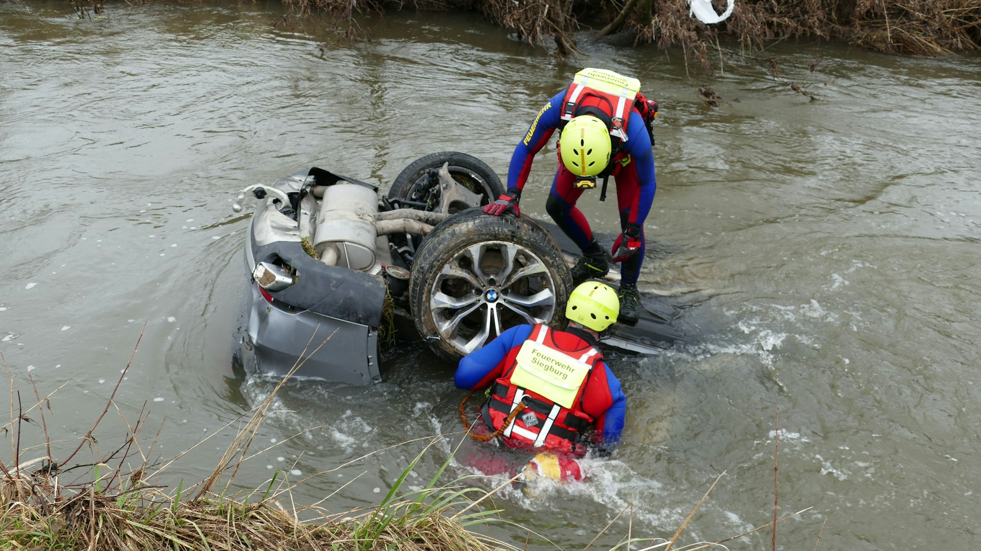 Rettungstaucher stehen an einem Fluss. Ein Auto schwimmt im Wasser.
