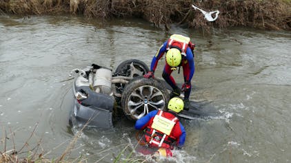 Rettungstaucher stehen an einem Fluss. Ein Auto schwimmt im Wasser.