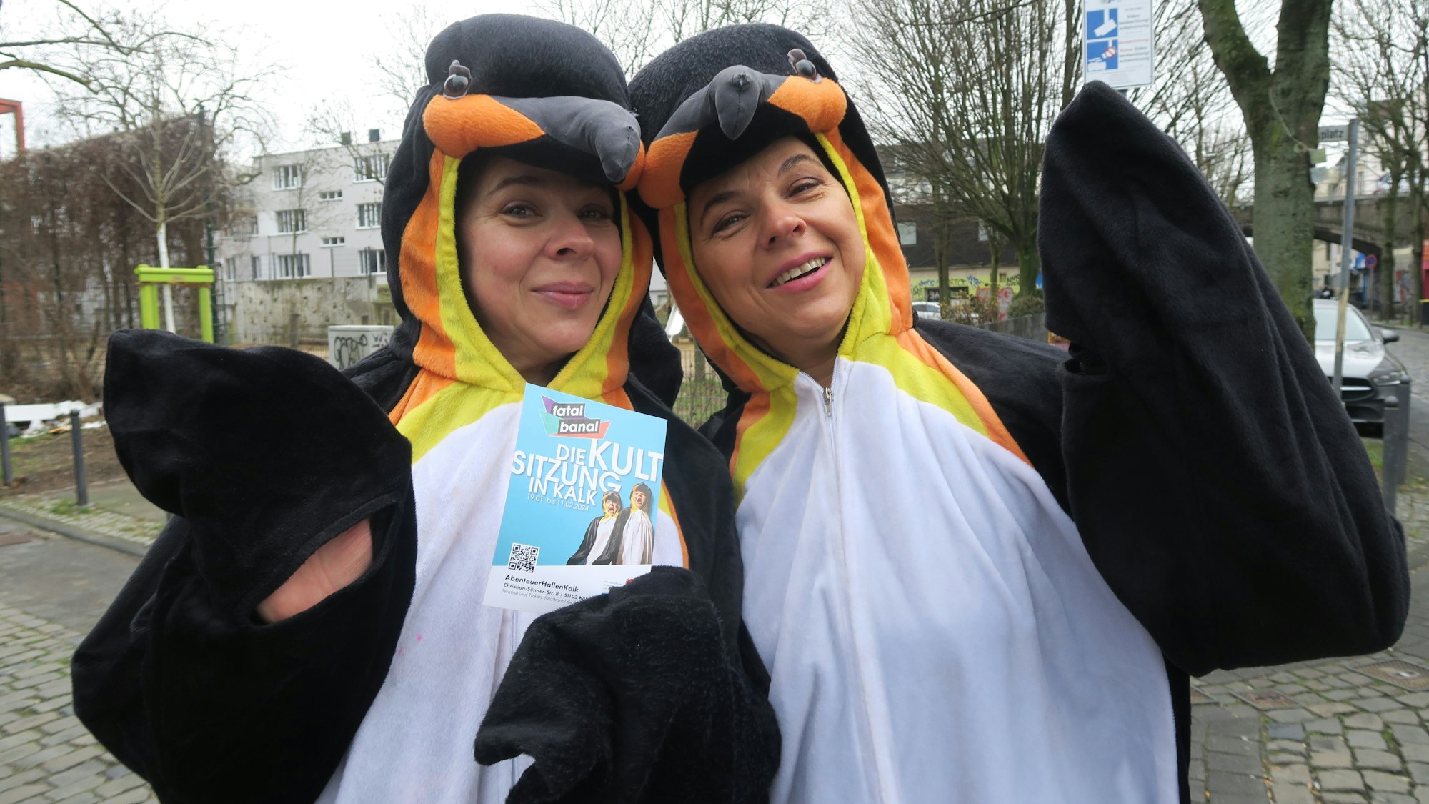 Zwei Frauen im Pinguin-Kostüm zeigen den Flyer der Kultsitzung Fatal Banal.