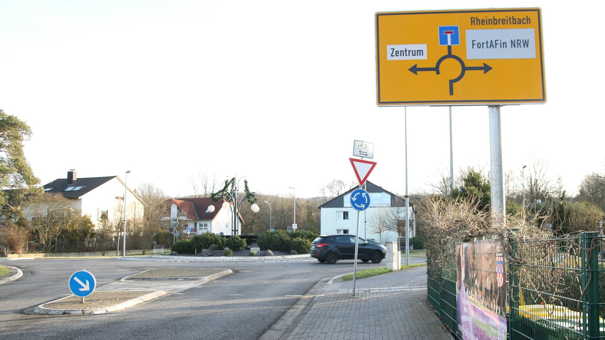 Ein Kreisverkehr, über den gerade ein blaues Auto fährt, und Schild mit der Wegweisung ins Zentrum oder nach Rheinbreitbach.