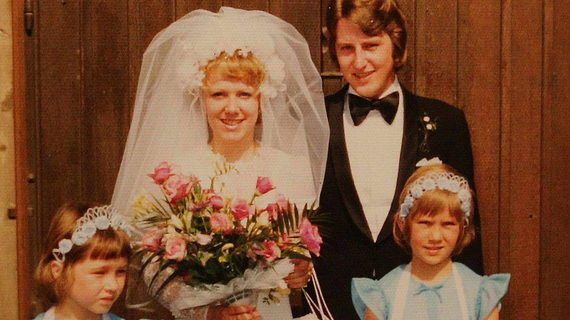 Die Braut im weißen Kleid mit Schleier, der Bräutigam mit Anzug und Fliege und mit dabei zwei Brautjungfern.